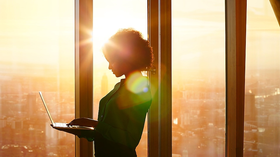 Woman standing using a laptop against a sunset behind windows in a building
